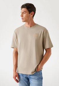 Beige bomulds t-shirt med rund halsudskæring, korte ærmer og et lille broderet logo på brystet. Casual pasform, blød tekstur.