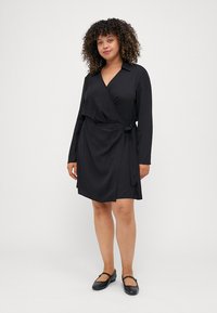 Abito avvolgente nero con colletto e maniche lunghe, caratterizzato da un dettaglio di legatura laterale. Tessuto liscio e design fino al ginocchio. Abbinato a scarpe basse.
