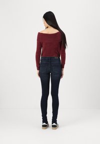 Pulover burgund cu umerii la vedere, realizat dintr-un tricot texturat, asociat cu blugi skinny de culoare albastru închis. Încălțămintea are accente negre și albe.