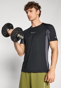 Svart och grå sporttröja med korta ärmar och ribbad textur, ihop med gröna shorts, håller en skivstång. Champion-logotyp synlig.