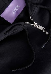 Fato de capuz preto com fecho zip metálico, cordão ajustável e etiqueta roxa com "JUXX." Tecido macio com uma textura suave.