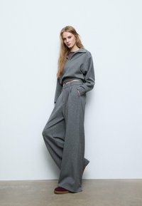 Szary sweter z kołnierzykiem polo, zestawiony z szerokimi szarymi spodniami z kontrastowym pasem. Model stoi na gładkiej podłodze.