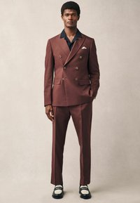 Brown double-breasted suit with gold buttons and black collar; paired with tailored pants and black shoes with white accents.