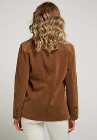 Veste en daim marron au design ajusté, dotée de manches longues et de trois boutons marron sur chaque poignet. Texture lisse et style minimaliste.