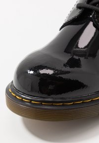 Schwarzer Lacklederschuh mit abgerundeter Spitze, gelber Naht entlang der Sohle und strukturierter brauner Gummilaufsohle.