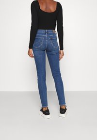 Jeans ajustados de color azul con cintura de altura media. Presenta dos bolsillos traseros y un diseño de pierna ajustada. Combinados con una camiseta negra de manga larga y zapatillas.