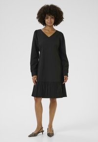 Robe noire avec un décolleté en V et des manches longues, présentant un ourlet plissé. Assortie à des escarpins à semelle zèbrée. Texture du tissu lisse.