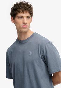 Camiseta de algodón gris con mangas cortas, cuello redondo y un pequeño logo bordado en el pecho izquierdo. Textura suave y ajuste casual.