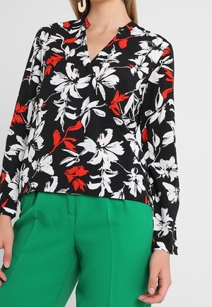 Blouse portefeuille noire à motif floral blanc et rouge, associée à un pantalon vert vif, portée par une personne debout sur un fond uni.