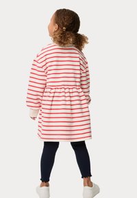 Gestreiftes Kleid in Weiß mit roten Streifen, mit einem taillierten Bund und lockeren Ärmeln, kombiniert mit navyblauen Leggings und weißen Schuhen.
