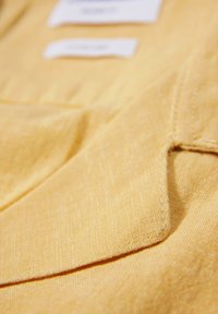 Tessuto giallo con una superficie texturizzata, presenta un angolo piegato e cuciture visibili. Comprende etichette per la cura in bianco nella parte superiore.