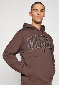 Sweatshirt castanho com um bolso canguru à frente, grande logotipo "NIKE" em cor contrastante, tecido texturizado e capuz com cordão ajustável.