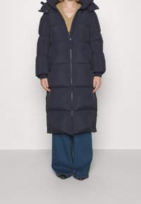 Langer Steppmantel in Dunkelblau mit Reißverschluss vorne, Kapuze und großen, gesteppte Abschnitten. Getragen über beigen Strickpullover und weiten Jeanshosen.