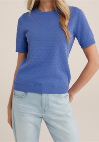 Blå stickad tröja med kort ärm som har strukturerade bubbelmönster, rundad halsringning och ribbade ärmslut och nederkant. Matchas med ljus denimjeans.