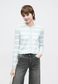 Světle modrý pletený kardigan s bílými pruhy, kulatým výstřihem a zapínáním na knoflíky. Kombinován s tmavě šedými širokými džínami.