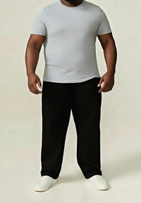 Camiseta gris de manga corta, pantalones negros holgados y zapatos blancos de tipo slip-on, llevados por una persona de pie contra un fondo neutro.