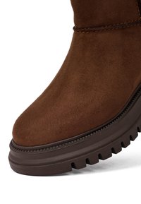 Bruine suède enkelboot met een afgeronde neus, gestructureerd oppervlak en een dikke, ribbelige rubberen zool. Bevat contrasterende bruine stikdetails.