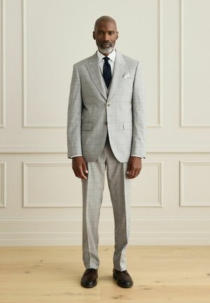 Man standing against paneled wall wearing light gray plaid suit, white shirt, dark tie, and dark shoes, facing forward with neutral expression.