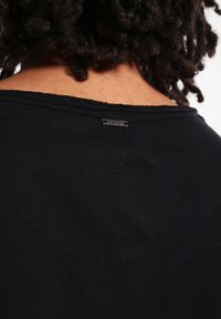 Zwarte katoenen t-shirt met een ronde halslijn, gladde textuur en een klein metalen label op de achterkant. Eenvoudig ontwerp zonder patronen.