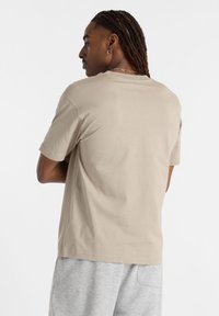 Tricou bej cu guler rotund, confecționat din bumbac neted, cu mâneci scurte și o croială lejeră. Spatele prezintă cusături minime și fără grafice.