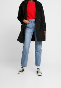 Sort tekstureret frakke med reverskrave, rød strikket sweater, lyseblå jeans med lige ben og sorte sneakers med hvide såler.