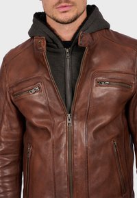 Bruine leren jas met een slim fit ontwerp. Voorzien van twee ritsborstzakken, een staande kraag en een grijze hoodie eronder.