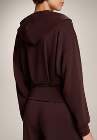 Brązowa bluza z kapturem, wykonana z miękkiego materiału, o luźnym kroju, obniżonych ramionach i ściągaczu w pasie. Gładki, minimalistyczny design.