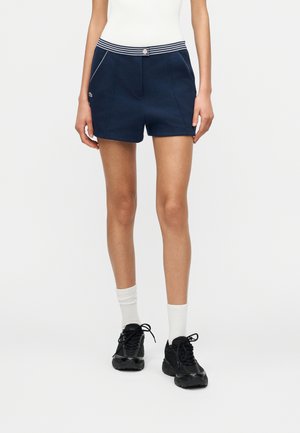 Vrouw draagt marineblauwe korte broek met wit gestreepte tailleband, wit hemd, witte sokken tot aan de scheen, en zwarte sneakers, staand tegen een witte achtergrond.