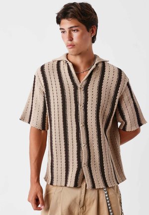 Young man wearing a beige and black striped short-sleeve knit shirt and beige pants, looking to the side with a neutral expression.