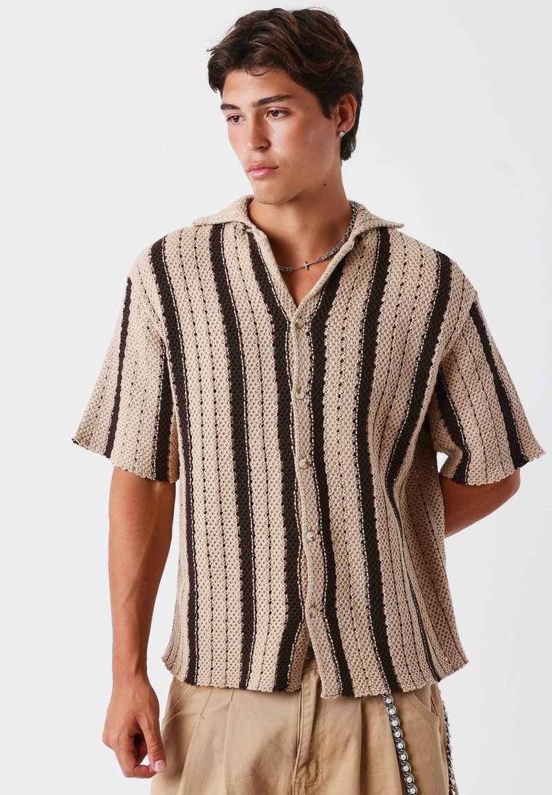 Jeune homme portant un pull en maille à manches courtes beige et noir à rayures et un pantalon beige, regardant sur le côté avec une expression neutre.