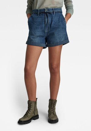 Shorts en denim avec des ourlets roulés, dotés d'un cordon de serrage et d'une fermeture à bouton ; couleur bleu profond avec une texture lisse, associés à des bottes de combat olive.