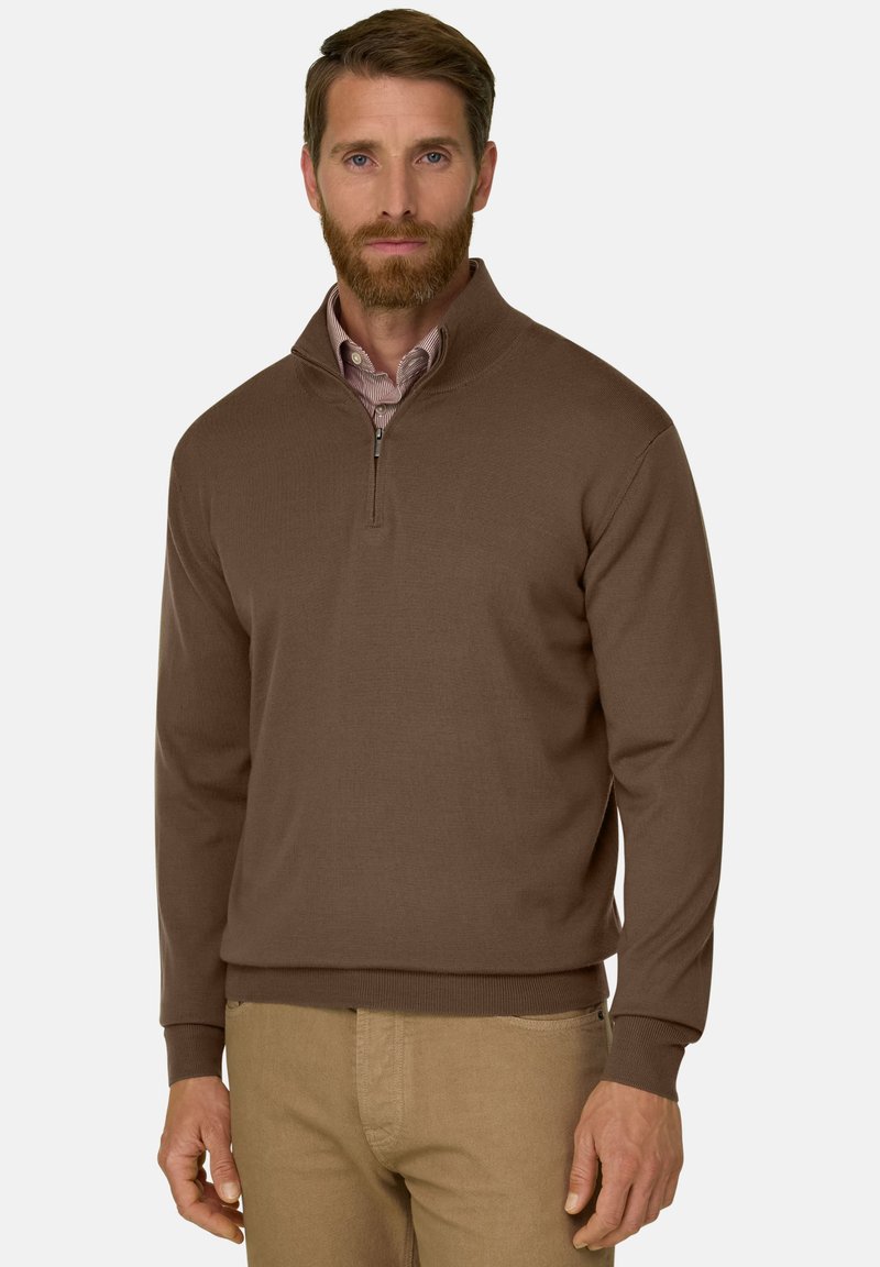 Homme avec barbe portant un pull marron à fermeture éclair jusqu’au quart, par-dessus une chemise rayée et un pantalon beige, debout devant un fond blanc uni.