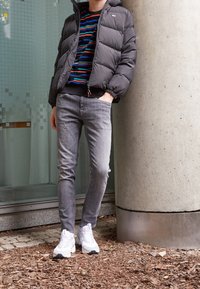 Šedá péřová bunda, vícebarevný pruhovaný svetr, šedé úzké džíny a bílé robustní tenisky. V pozadí je beton a zeleň.