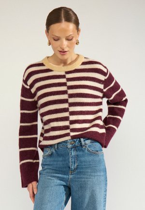 Vrouw draagt een bordeauxrode en beige gestreepte pluizige trui met blauwe high-waist jeans, kijkt naar beneden met één hand op haar heup.