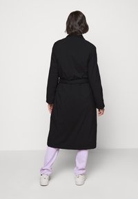 Schwarzer langer Mantel mit Gürtel, mit Reverskragen und langen Ärmeln. Kombiniert mit lila Hosen und weißen Turnschuhen. Glatte Textur.