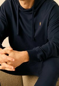 Granatowa bluza z kapturem, wykonana z miękkiego materiału, z kieszenią kangurką i sznurkiem do regulacji, z małym złotym logo na klatce piersiowej.