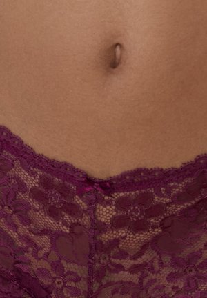 Nahaufnahme des Mittelbereichs, die nackte Haut und lila Spitzenunterwäsche mit floralem Muster sowie einer kleinen Schleife in der Mitte zeigt.