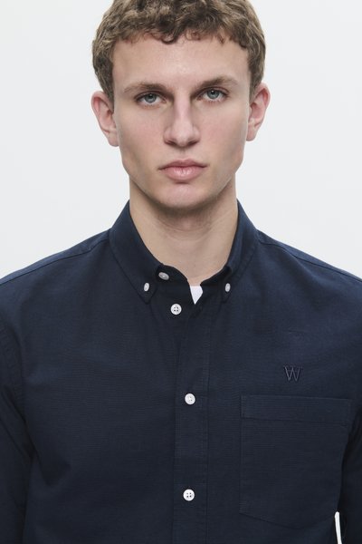 Jeune homme avec des cheveux bouclés courts portant une chemise à boutons bleu foncé avec une poche poitrine sur un fond blanc uni.