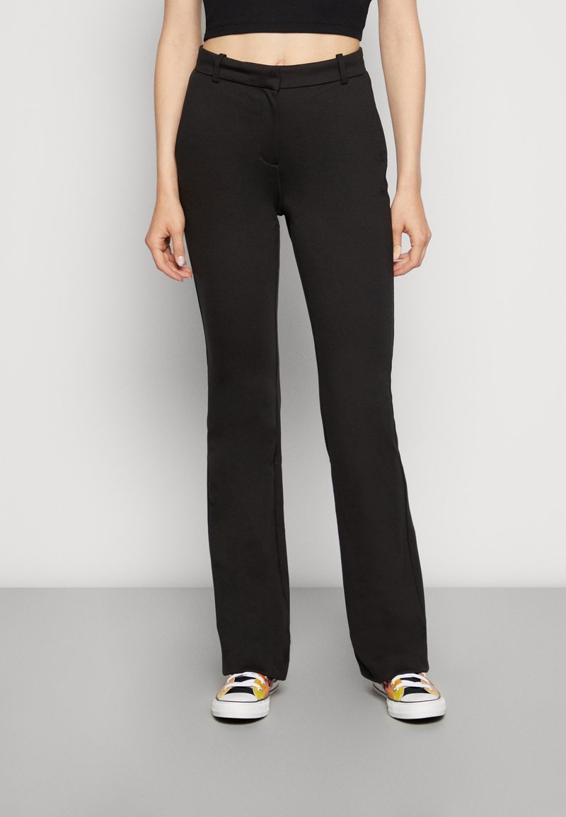 Femme portant un pantalon noir taille haute à jambes droites, associé à des baskets colorées et un haut court noir, debout sur un fond uni.