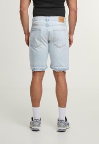 Világoskék farmer rövidnadrág, rojtos szegéllyel, öt zsebes kialakítással és bézs bőrpáttal a hátsó derékszíjon. Fehér zoknival és sportcipővel párosítva.