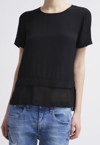 Svart kortärmad blus med lagerdelsprint, tillverkad av lättviktig tyg. Matchad med blå slitna jeans. Slät textur och enkel design.