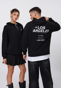Sudaderas negras con bolsillos frontales y cordones, con el texto "LOS ÁNGELES" en la parte posterior, combinadas con una falda negra con pliegues y jeans oscuros.