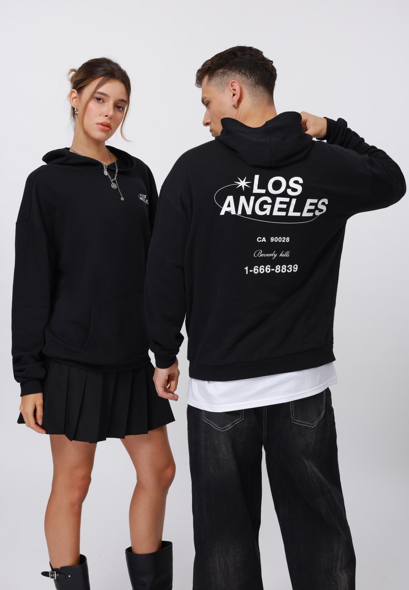 Sudaderas negras con bolsillos frontales y cordones, con el texto "LOS ÁNGELES" en la parte posterior, combinadas con una falda negra con pliegues y jeans oscuros.