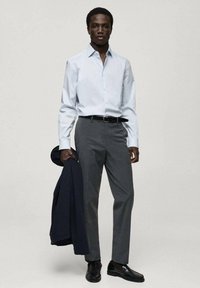 Cămașă bleu deschis, pantaloni gri și pantofi negri; ținând un sacou închis la culoare. Material neted, croi ajustat și design clasic.