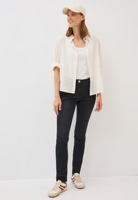 Camisa blanca ligera y transparente de botones sobre una camiseta blanca de tirantes, combinada con jeans oscuros ajustados y zapatillas crema con acentos burdeos.