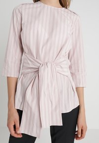 Blouse rose clair à manches longues avec des rayures blanches verticales, à nouer à la taille, avec un col bateau. Texture de tissu lisse.