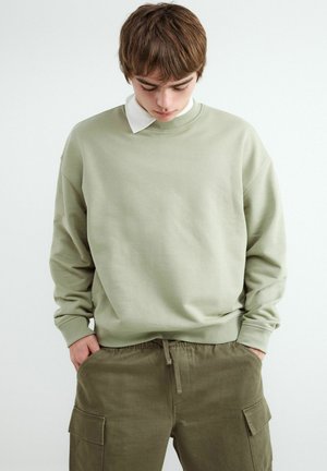 Jong persoon met kort bruin haar die naar beneden kijkt, draagt een lichtgroene trui over een wit overhemd met kraag en olijfgroene cargobroek.