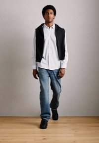 Mann, der drinnen geht, trägt ein weißes Hemd, einen schwarzen Hoodie über den Schultern, blaue Jeans und schwarze Sneakers auf einem Holzboden.
