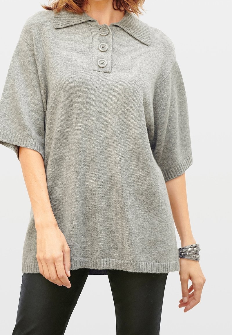 Grauer Strickpullover mit kragenförmigem Halsauschnitt, drei großen Knöpfen, kurzen Ärmeln und geripptem Saum, getragen über schwarzen Hosen.