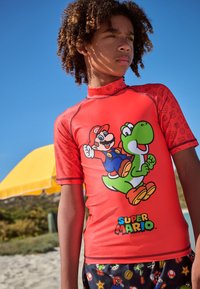 Red short-sleeve rash guard featuring Super Mario and Yoshi graphics. Smooth texture, high collar, with colorful patterned shorts.